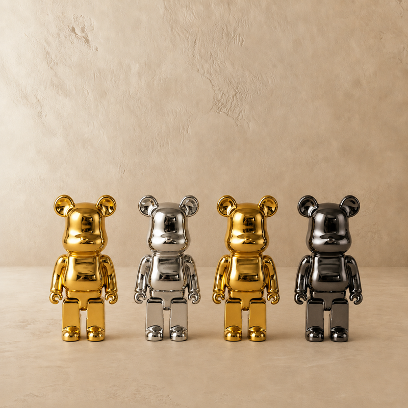 Four metallic bear-shaped collectible figures standing in a row on a beige surface against a textured beige wall. Two figures are shiny gold and two are metallic silver and dark gray.