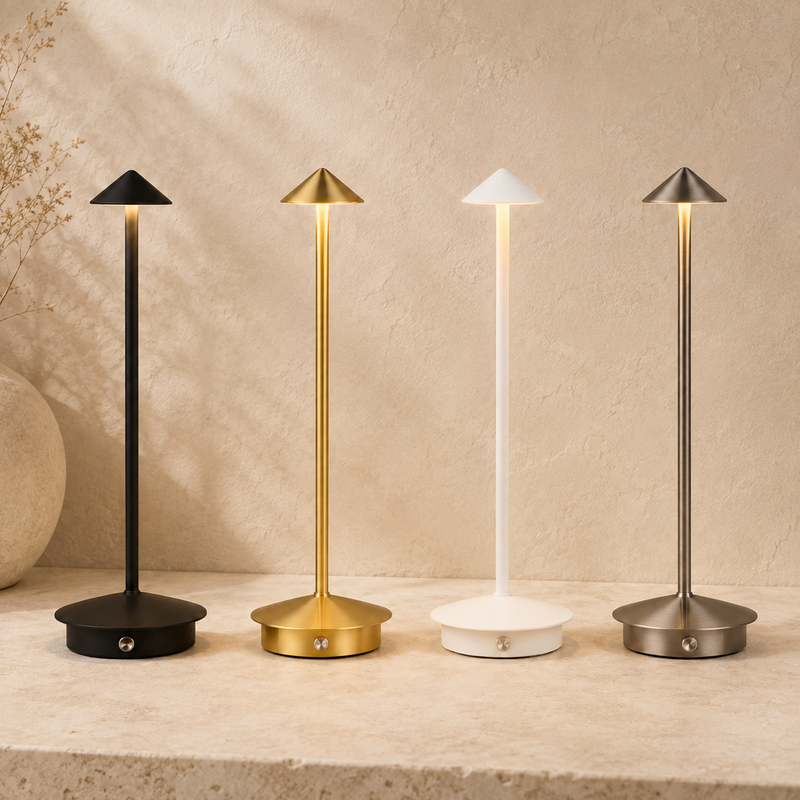 Four modern minimalist table lamps with slim stems and pyramid-shaped shades are lined up on a stone surface against a textured beige wall. The lamps come in matte black, brushed gold, white, and gunmetal gray finishes, all illuminated to show warm light.
